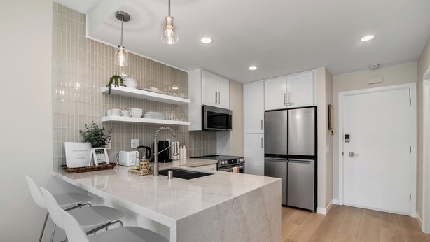 A modern kitchen with white cabinetry, a stainless steel refrigerator, a microwave, and a sink. There are two white bar stools and a hanging light fixture. - Condo on property at LaCosta Golf Resort & Spa with Pool 694 (Carlsbad)