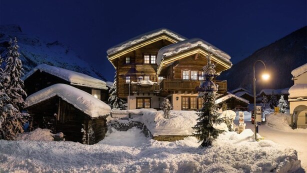 Eine große Holzhütte mit mehreren Balkonen, umgeben von schneebedeckten Bäumen und einer Bergkulisse.