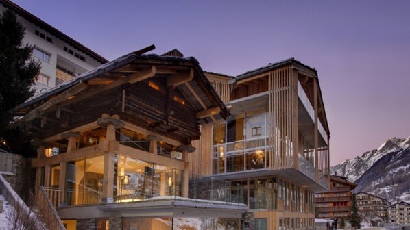Un moderno edificio de estación de esquí con amplios ventanales de vidrio y arquitectura de madera, rodeado de montañas nevadas.