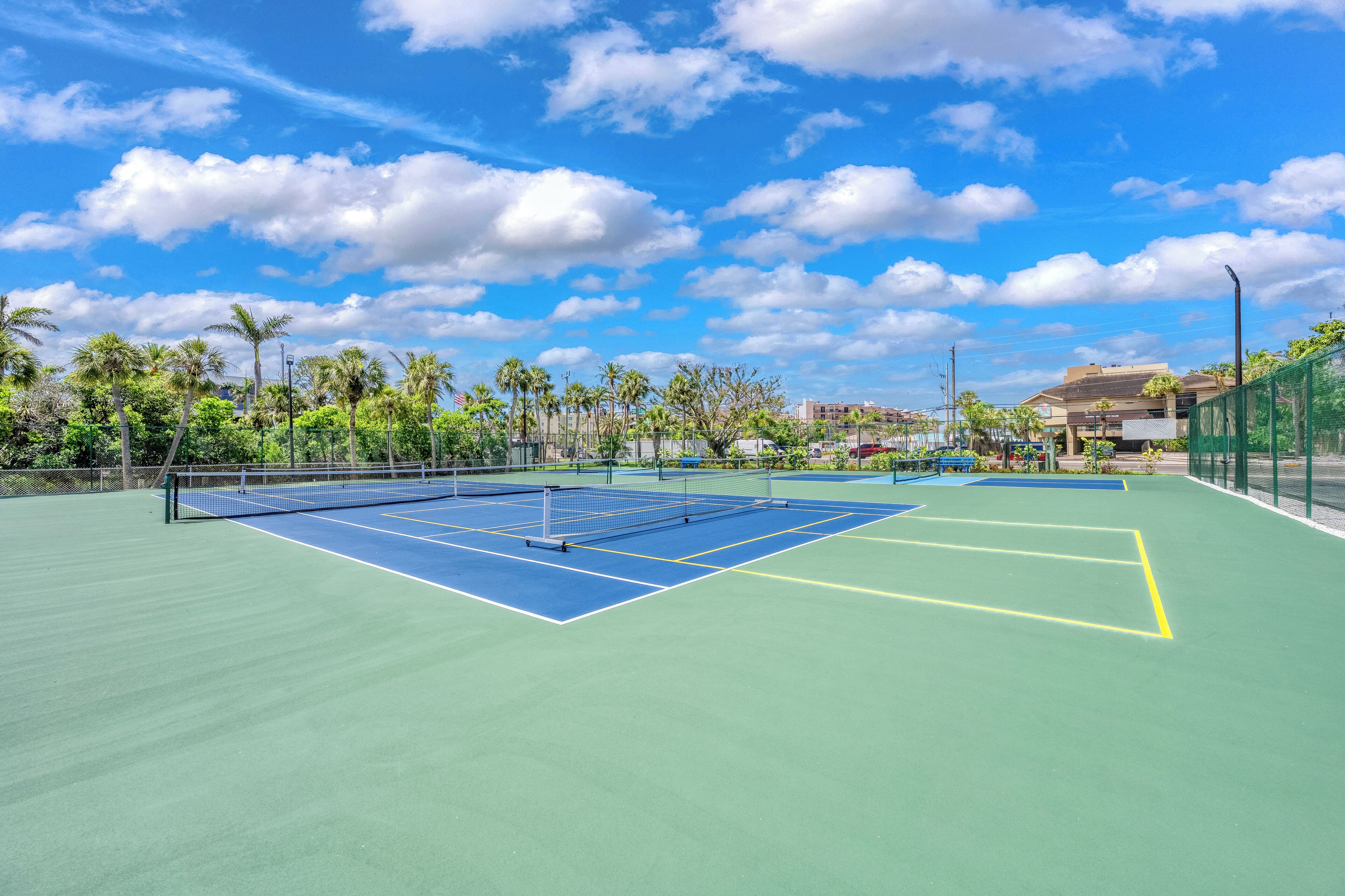 An outdoor tennis court with a net, surrounded by palm trees and a clear sky.