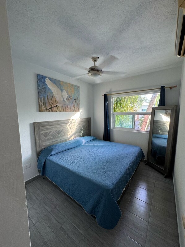 Une chambre à coucher avec un lit, un ventilateur de plafond, une fenêtre avec des rideaux et un tableau au mur.