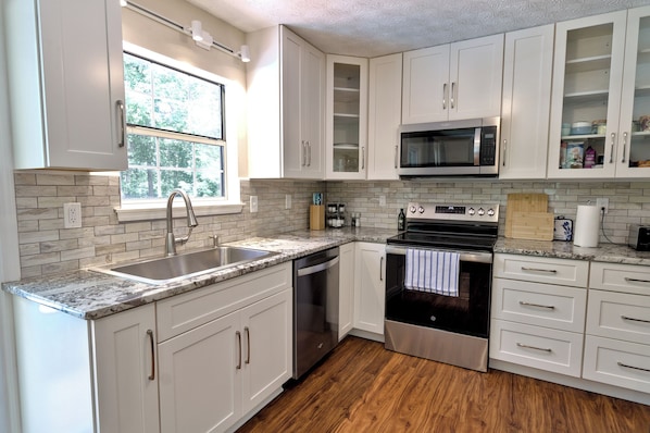 A modern kitchen with white cabinets, a stainless steel dishwasher, an oven, a microwave, and a window providing natural light.
