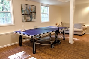 A ping pong table in a room with wooden flooring, two windows, a couch, and a painting on the wall.