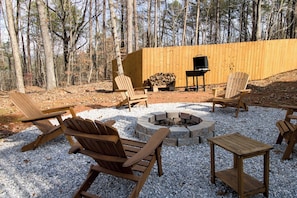 A backyard with a fire pit, wooden chairs, a table, and a grill.