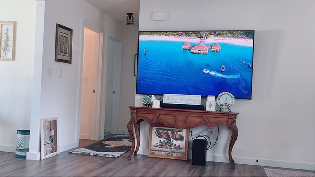 A living room with a large flat-screen TV mounted on the wall, a wooden console table with decorative items, and a framed painting below the TV. - New Home • 8 Miles to LBI .BBQ. Family friendly.  (Stafford Township)