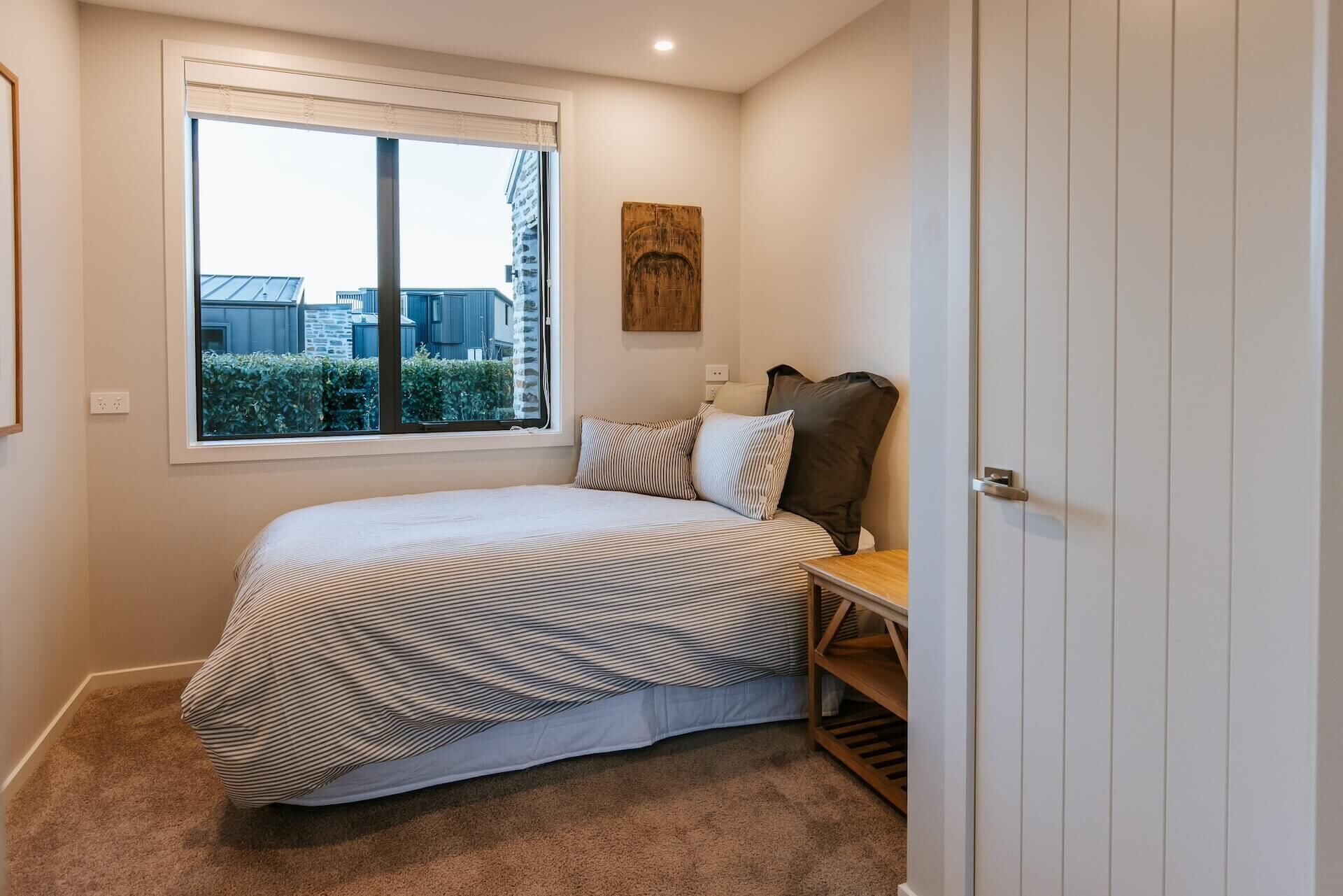 A bedroom with a large window, a bed with striped bedding, a wooden headboard, and a small wooden table.