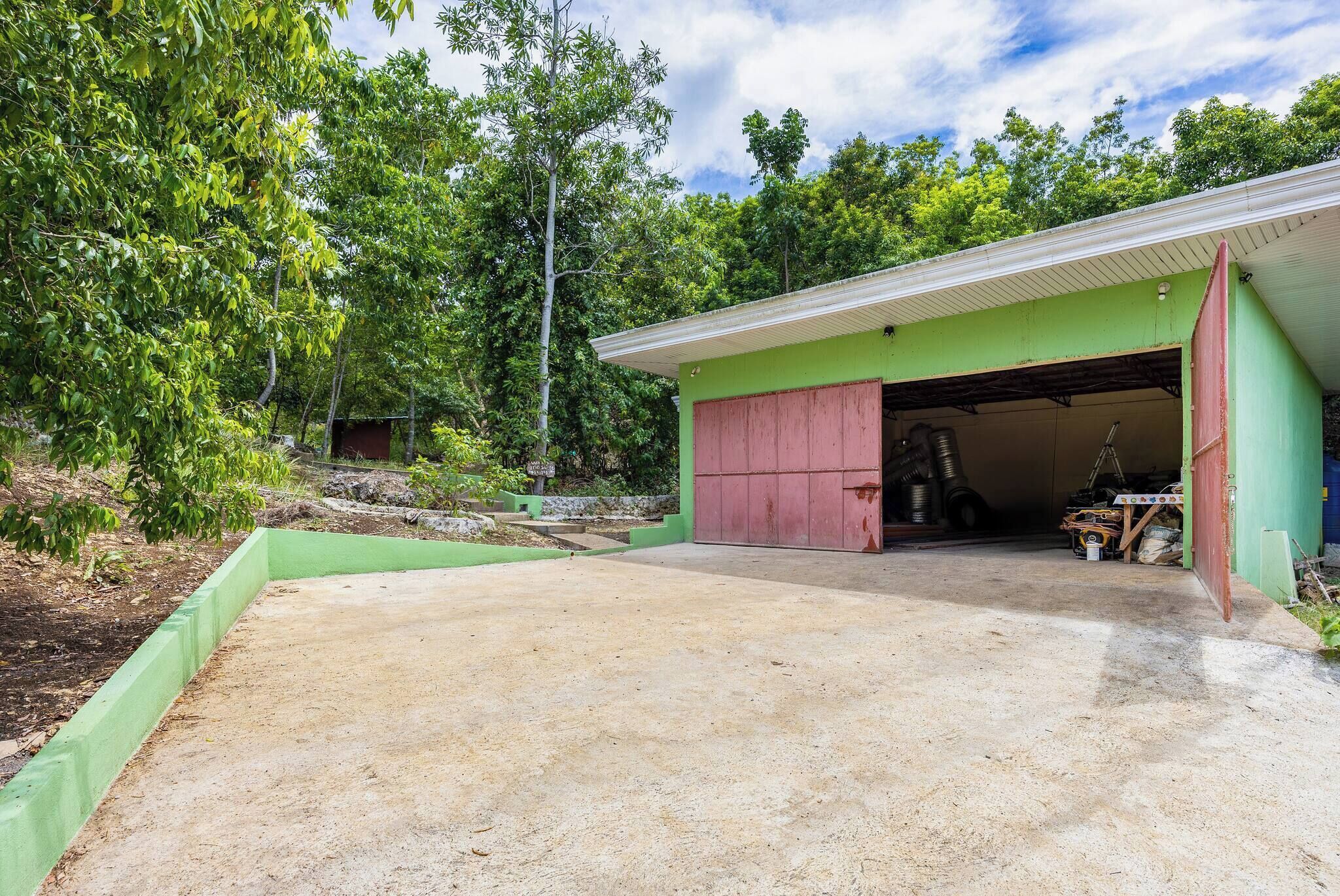 A garage with a green exterior and a red door, surrounded by trees and a dirt driveway.