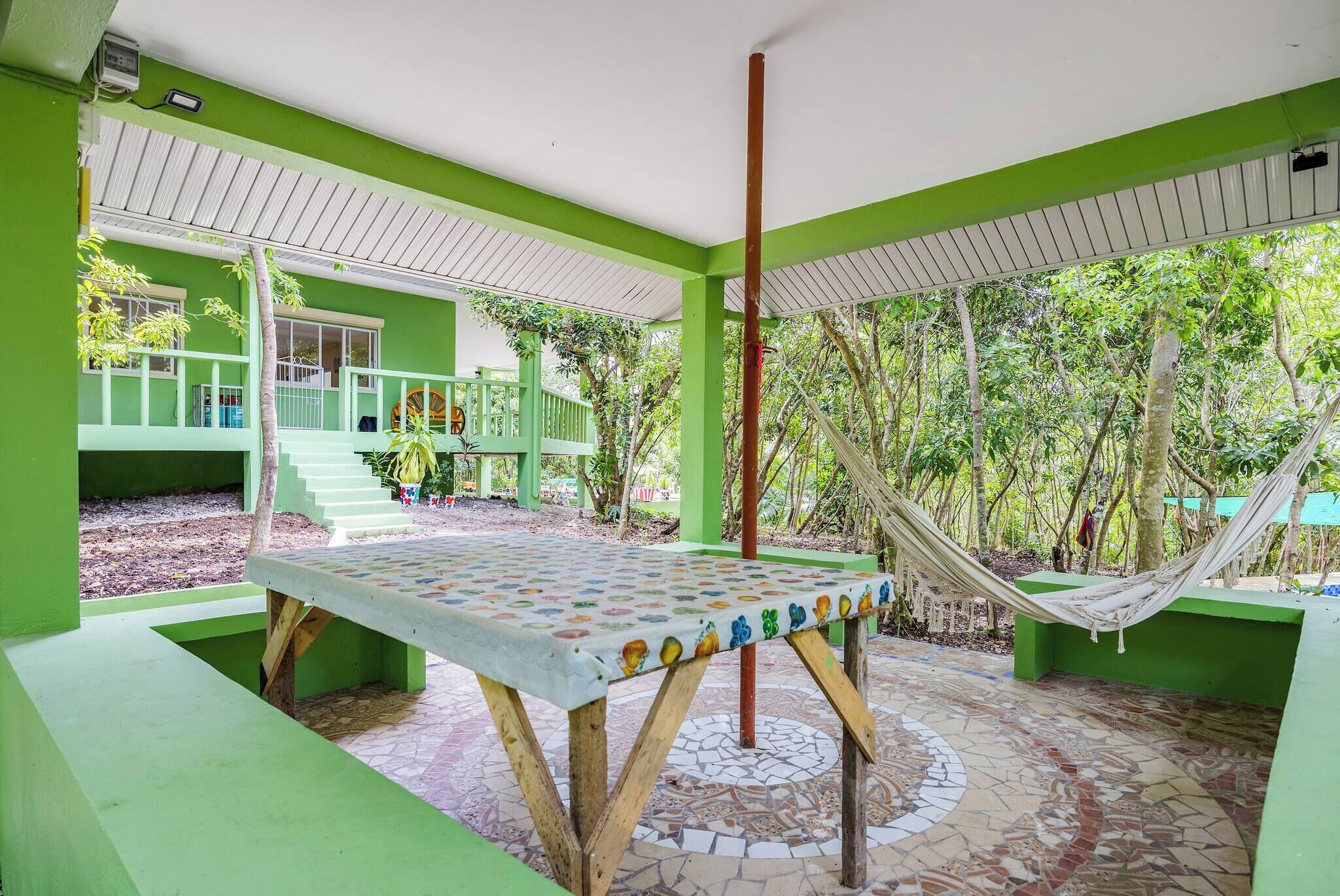 A green-roofed outdoor area with a tiled floor, a hammock, and a table with a colorful pattern.