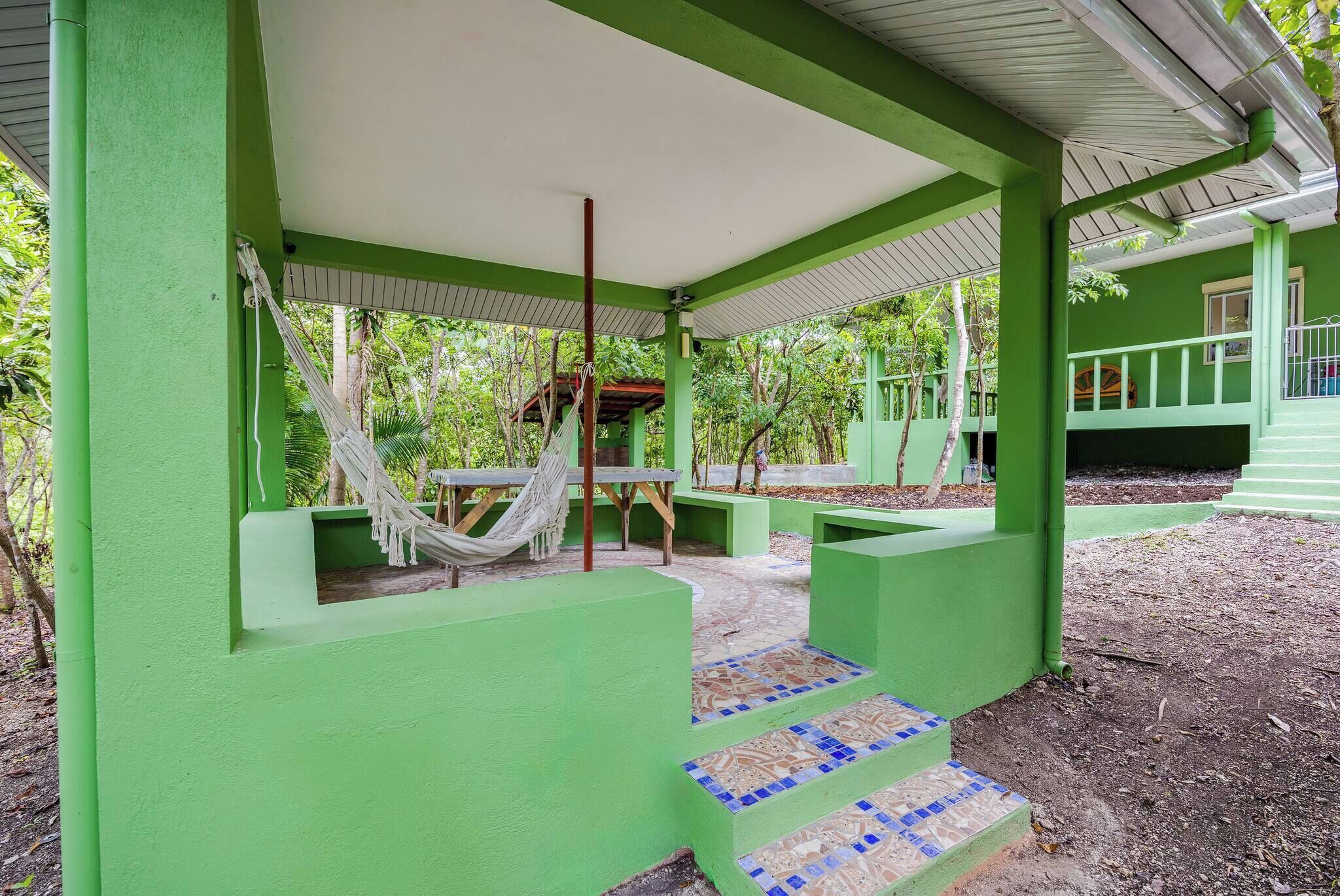 Un espacio al aire libre con techo verde, una hamaca, escalones y piso de baldosas.