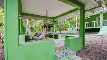 Un espacio al aire libre con techo verde, una hamaca, escalones y piso de baldosas.