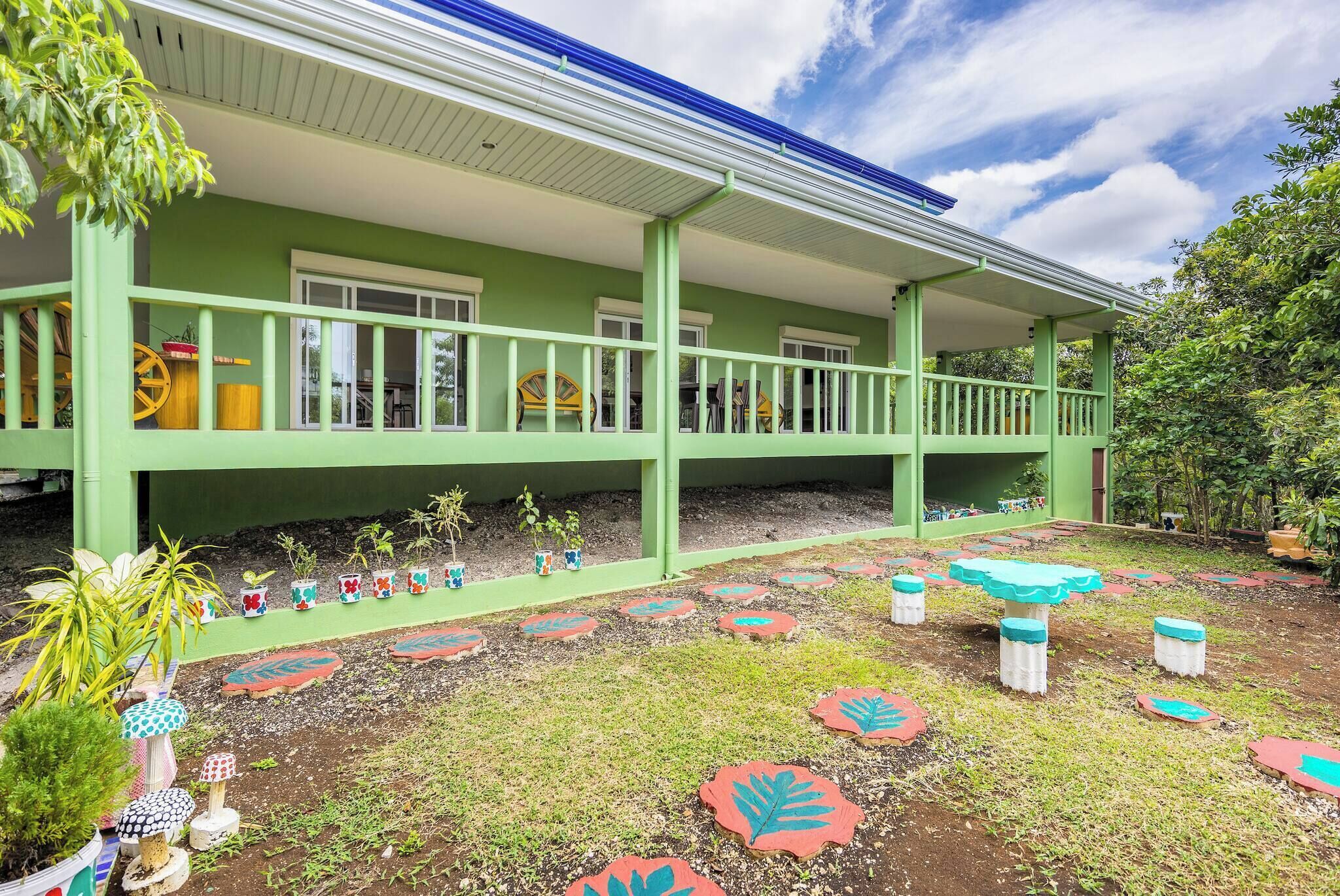 A green building with a covered porch, potted plants, and a garden with decorative tiles.