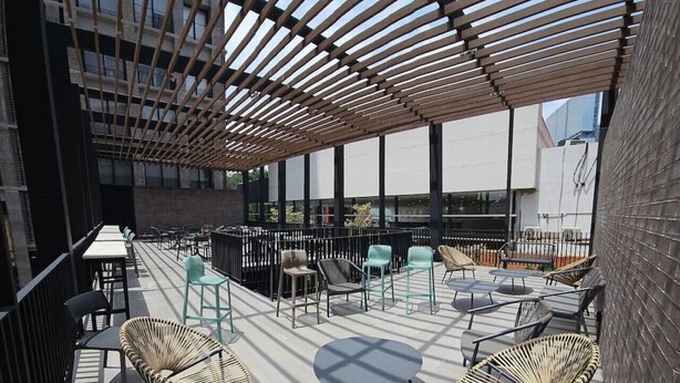 An outdoor seating area with wicker chairs and tables, a wooden pergola, and a view of a modern building. - New apartment with beautiful views in Lomas del Country (Guadalajara)
