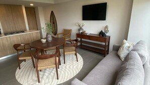 Un salon moderne avec un canapé gris, une table à manger avec des chaises, un téléviseur fixé au mur et un coin cuisine à l’arrière-plan.