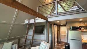 A loft-style room with a wooden floor, a sofa, a striped chair, a ladder, a kitchen area with a refrigerator, and a mezzanine level with a mattress.