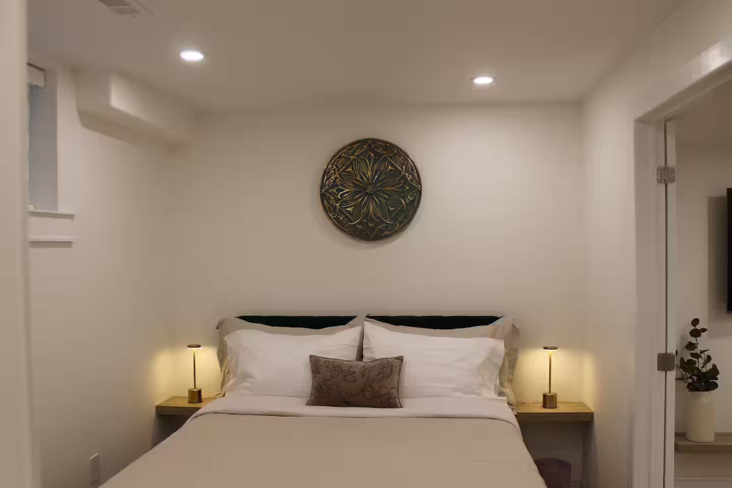 A bedroom with a bed, two bedside tables, a wall-mounted decorative piece, and a door leading to another room.