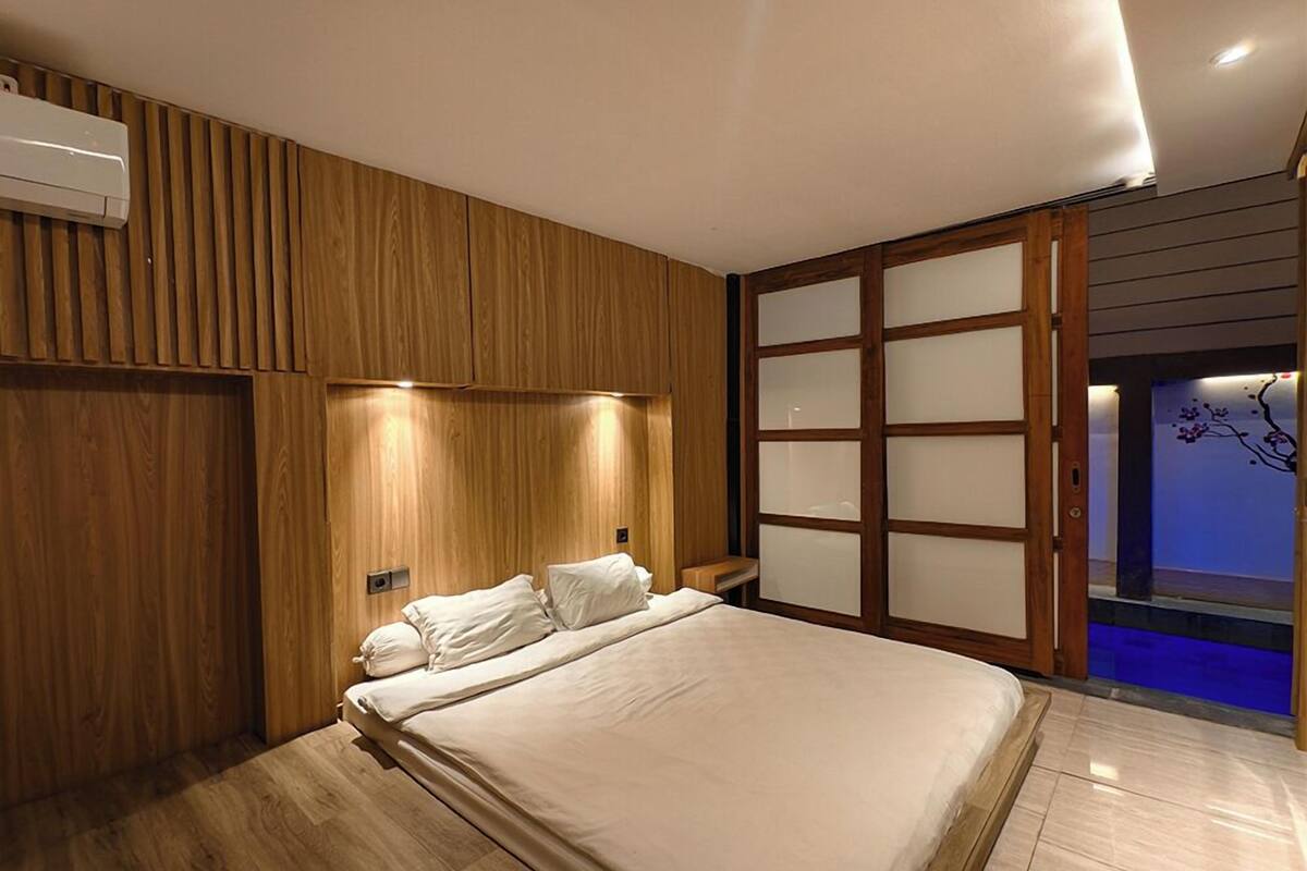 A modern bedroom with a wooden panel wall, a bed with white linens, and a sliding glass door leading to a blue-lit area.
