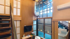 A modern living room with a TV, wooden paneling, a large window, and a staircase. - Ohana Suites Yogyakarta (Yogyakarta)