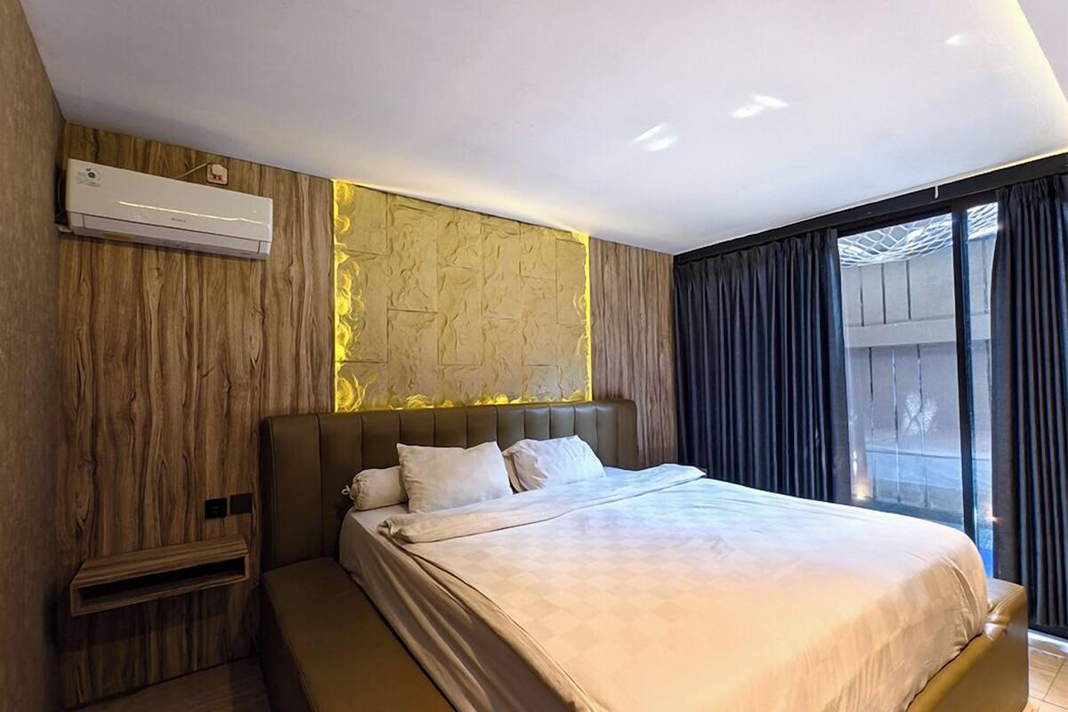 A modern bedroom with a large bed, a built-in air conditioning unit, a wooden accent wall, and a view through large windows.