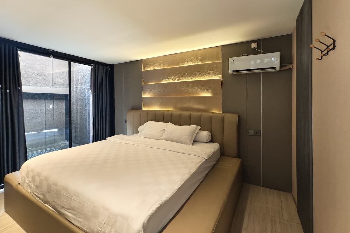 A modern hotel room with a large bed, a built-in wall-mounted air conditioning unit, and a view of the outdoors through a large window.