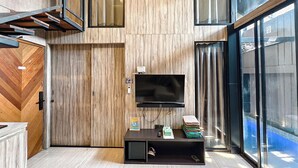 A modern living room with a wooden wall panel, a flat-screen TV mounted above a dark entertainment unit, and a glass door leading to a pool area. - Ohana Suites Yogyakarta (Yogyakarta)