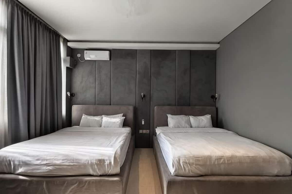 A hotel room with two beds, a wooden headboard, a window with curtains, and a wall-mounted air conditioning unit.