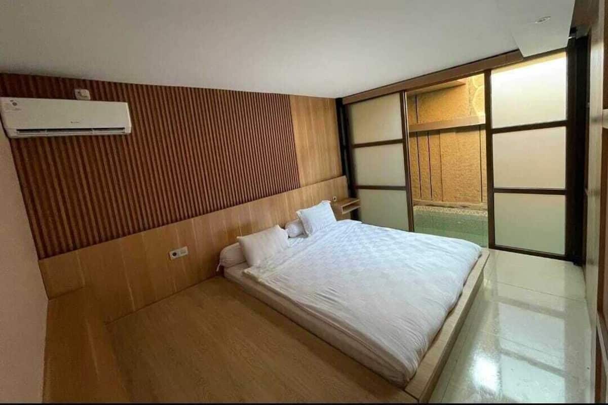 A hotel room with a wooden headboard, a bed with white bedding, an air conditioning unit, and a sliding glass door leading to a balcony.