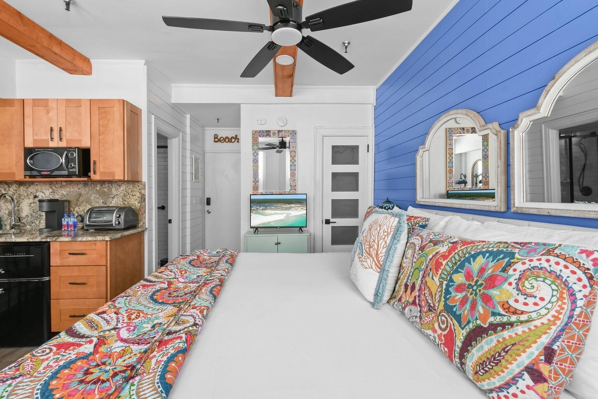 A bedroom with a bed, a TV, a ceiling fan, and a kitchenette.