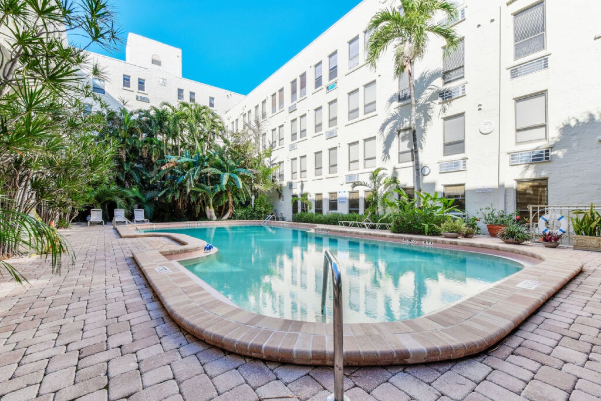 A swimming pool with a tiled deck, surrounded by lush greenery and a multi-story building with balconies.