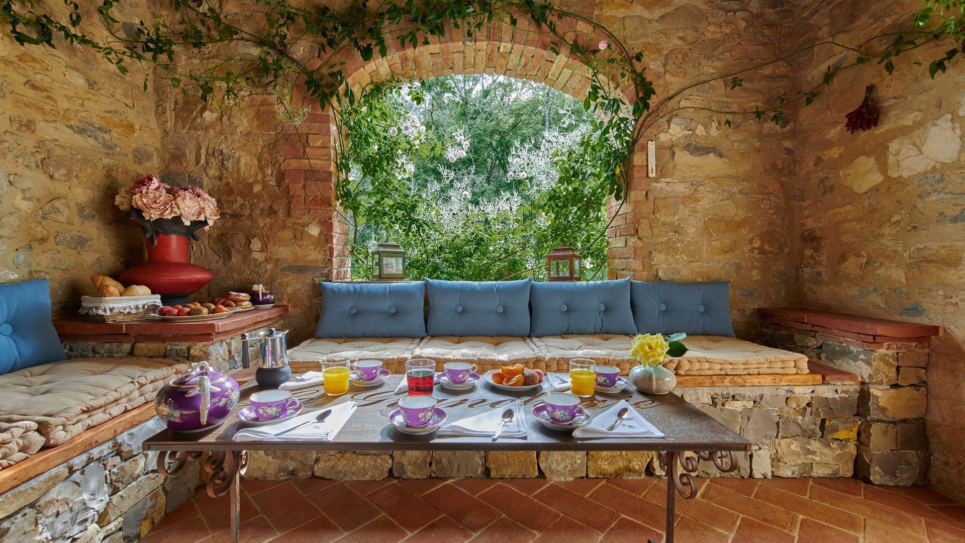 A rustic stone room with a bench, a table set for breakfast, and a view through a brick archway.