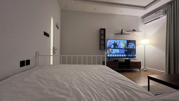 Un dormitorio con cama, televisor sobre un mueble bajo, mesita de noche, lámpara y aire acondicionado.