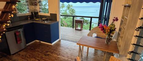 A cozy kitchen with a view of the ocean, a dining area with wicker furniture, and a wooden ceiling.