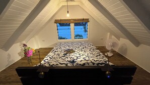 Un dormitorio con techo inclinado, una cama con colcha estampada, una ventana con persianas, un ventilador de pie y un florero con flores sobre una mesa de madera.