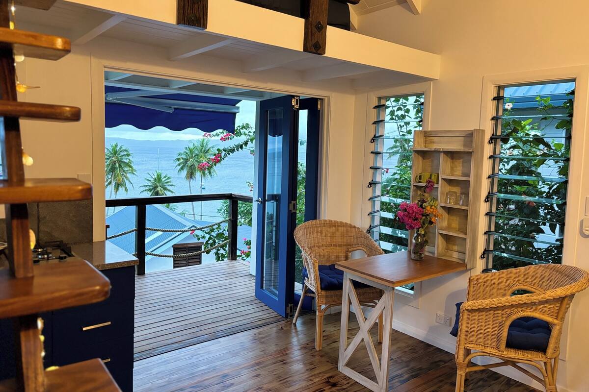 Habitació amb sòl de fusta, una cadira de vimetla, una taula petita i una prestatgeria. Es veuen palmeres i l'oceà a través d'una porta de vidre.