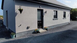 Un bâtiment d’un seul étage, à l’extérieur gris, avec une porte noire et un petit porche agrémenté de plantes en pot.