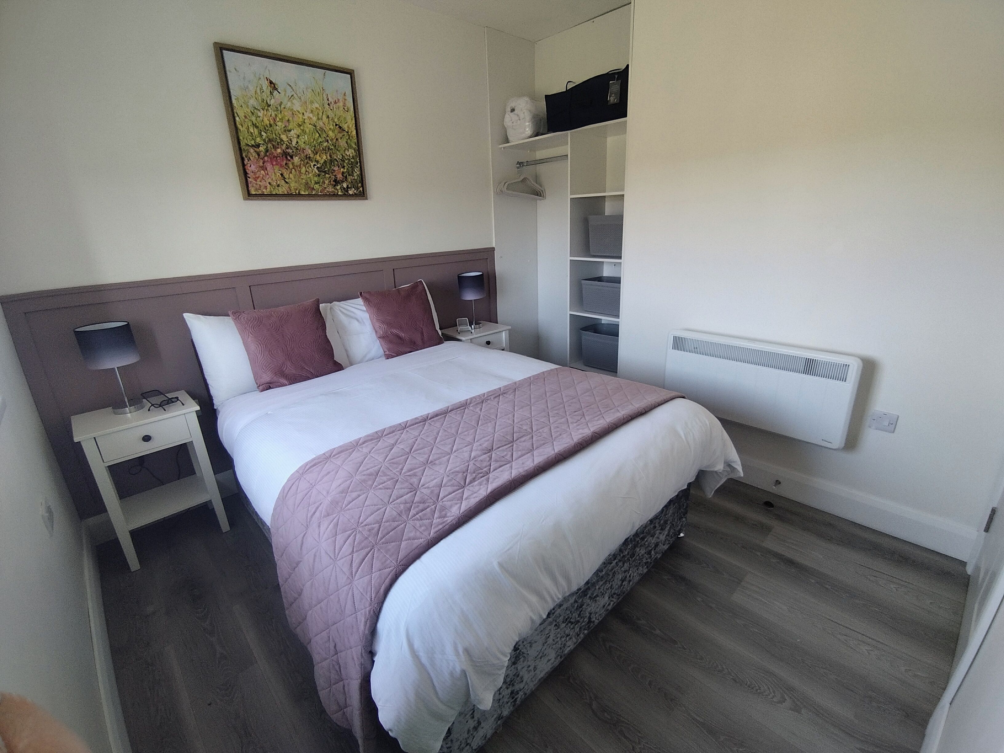 A neatly arranged bedroom with a bed, bedside tables, a nightstand, a radiator, and a wall-mounted shelf with storage baskets.