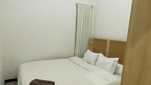 A single bed with a wooden headboard, white bedding, and a brown towel on the footboard. A window with white curtains is visible, and a white air conditioning unit is mounted on the wall. - Omah Kulon Malioboro (Kasihan)