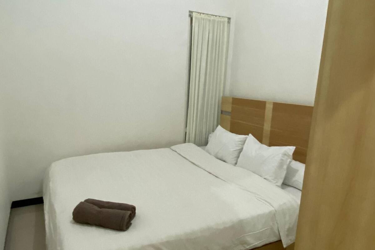 Een eenpersoonsbed met houten hoofdeinde, wit beddengoed en een bruine handdoek aan het voeteneinde. Een raam met witte gordijnen is zichtbaar, en een witte airconditioningunit is aan de muur bevestigd.