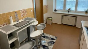 Une petite cuisine équipée d’un évier, de deux tabourets, d’une fenêtre avec des stores et d’un radiateur.