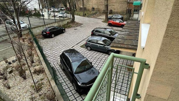 Un espace de stationnement où plusieurs voitures sont garées en rangées, une clôture métallique verte et un bâtiment en arrière-plan.