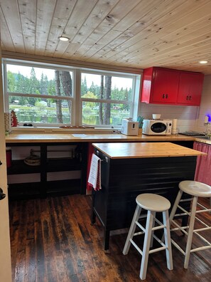 A kitchen with a wooden ceiling, a large window with a view of trees and water, red cabinets, a microwave, and two white stools.