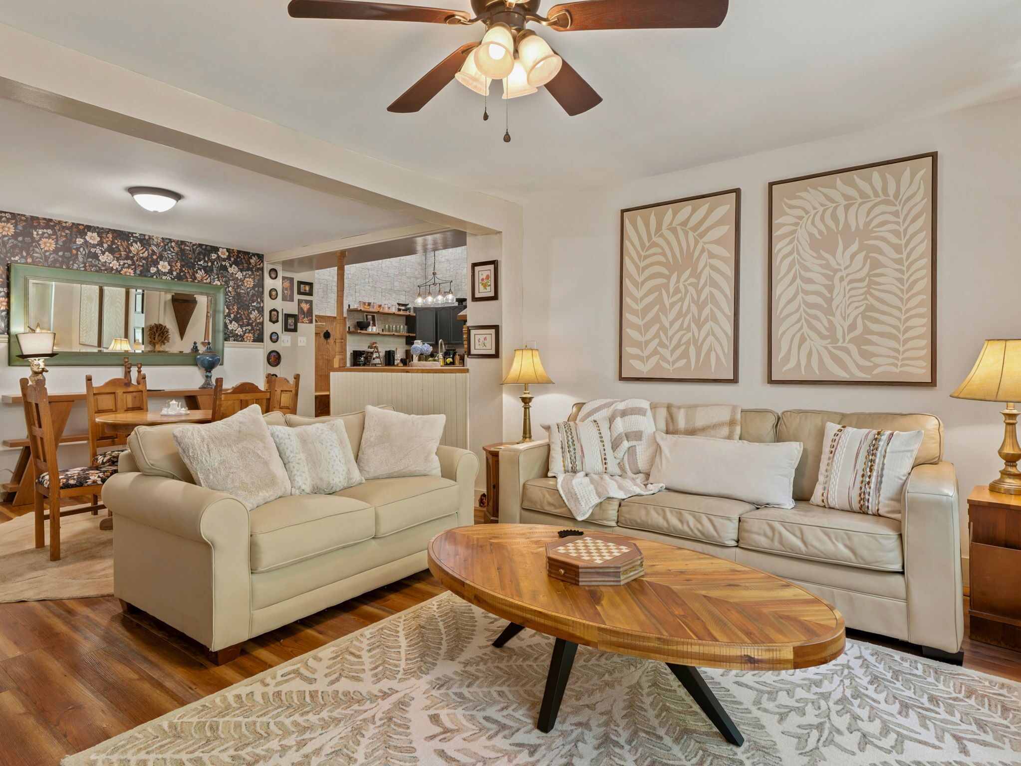 A living room with two sofas, a coffee table, a ceiling fan, and a kitchen area in the background.