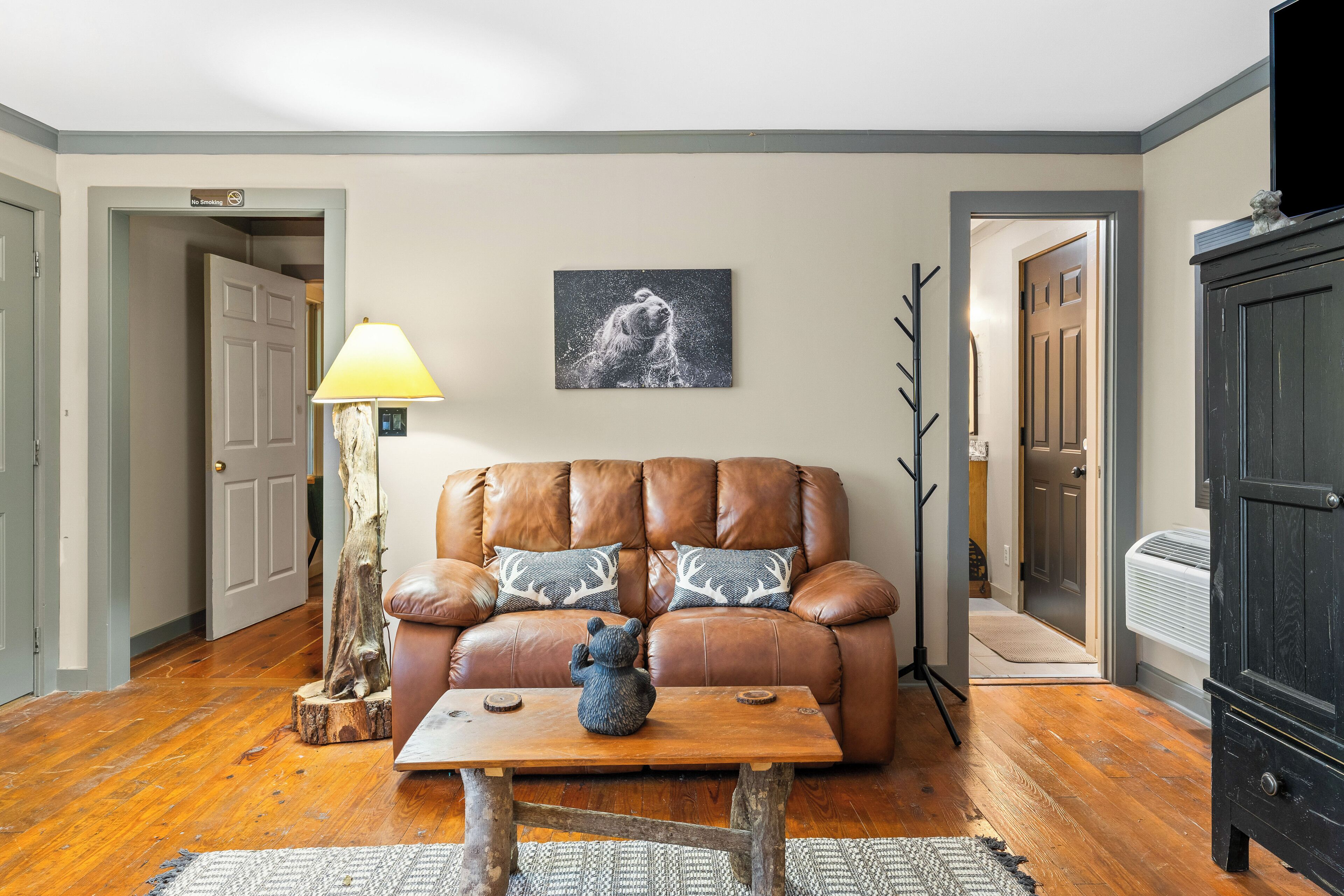 Un salon avec un canapé en cuir marron, deux coussins décoratifs à motifs, une table basse en bois ornée d’une sculpture d’ours, un tableau accroché au mur et une lampe.