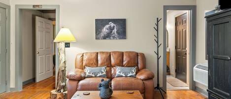 A living room with a brown leather sofa, two patterned throw pillows, a wooden coffee table with a bear sculpture, a wall-mounted picture, and a lamp.
