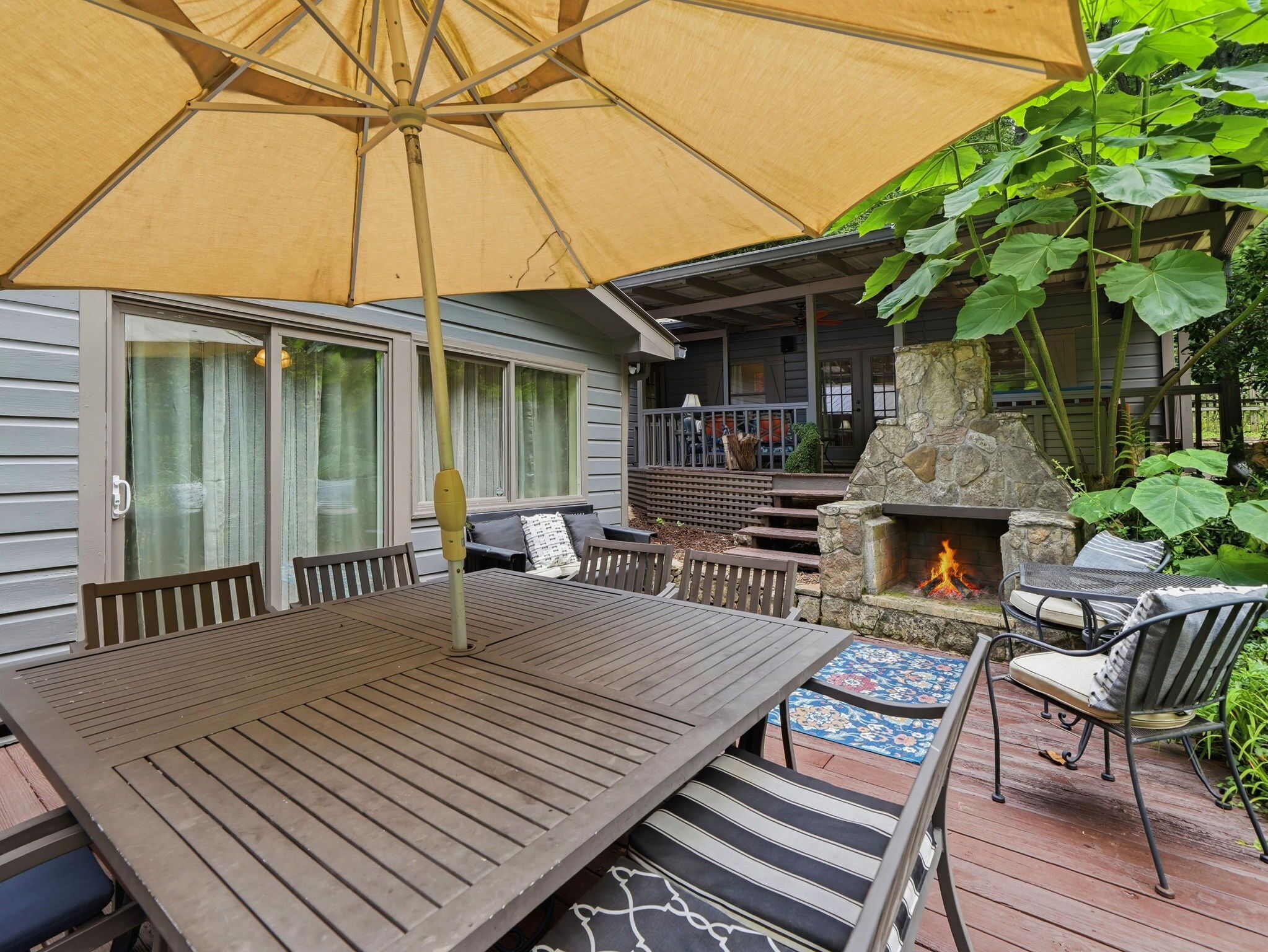 An outdoor patio with a wooden table and chairs, a stone fireplace, and a large umbrella providing shade.
