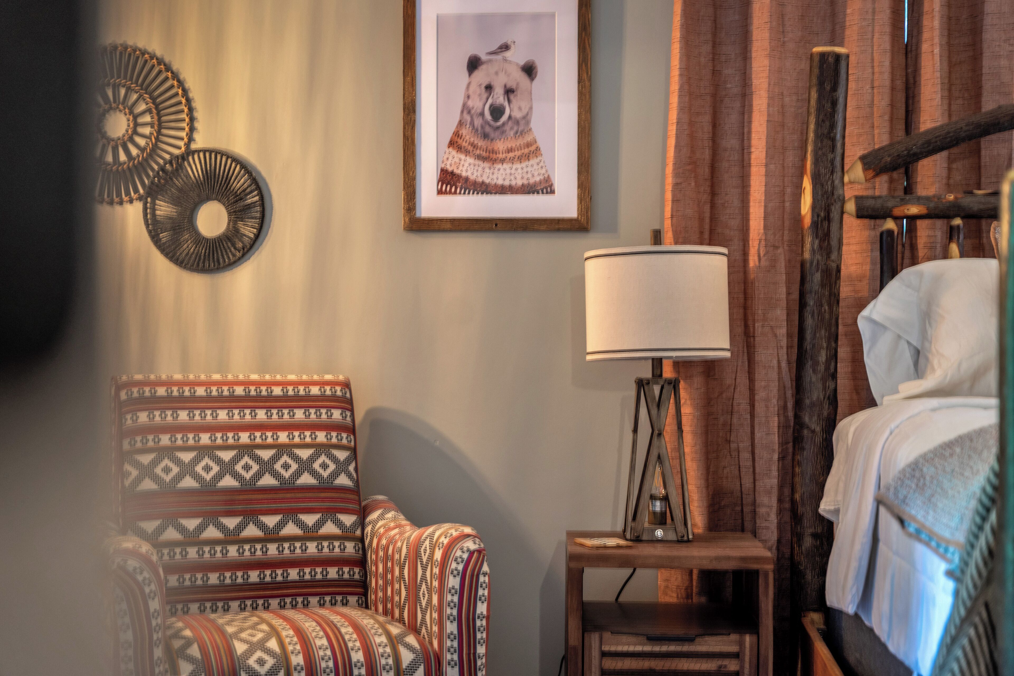 Een kamer met een gestreept fauteuil, een nachtkastje met een lamp, een lijst met een dierenafbeelding en een bed met een houten hoofdeinde.