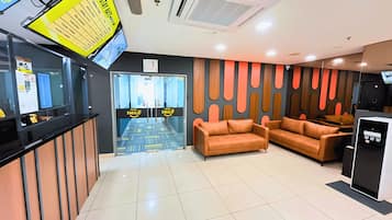 Uma área de recepção de hotel com um painel digital, balcão de recepção, espaço de estar com sofás e uma máquina de café.
