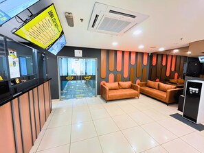 A hotel reception area with a digital display, a reception desk, a seating area with sofas, and a coffee machine.