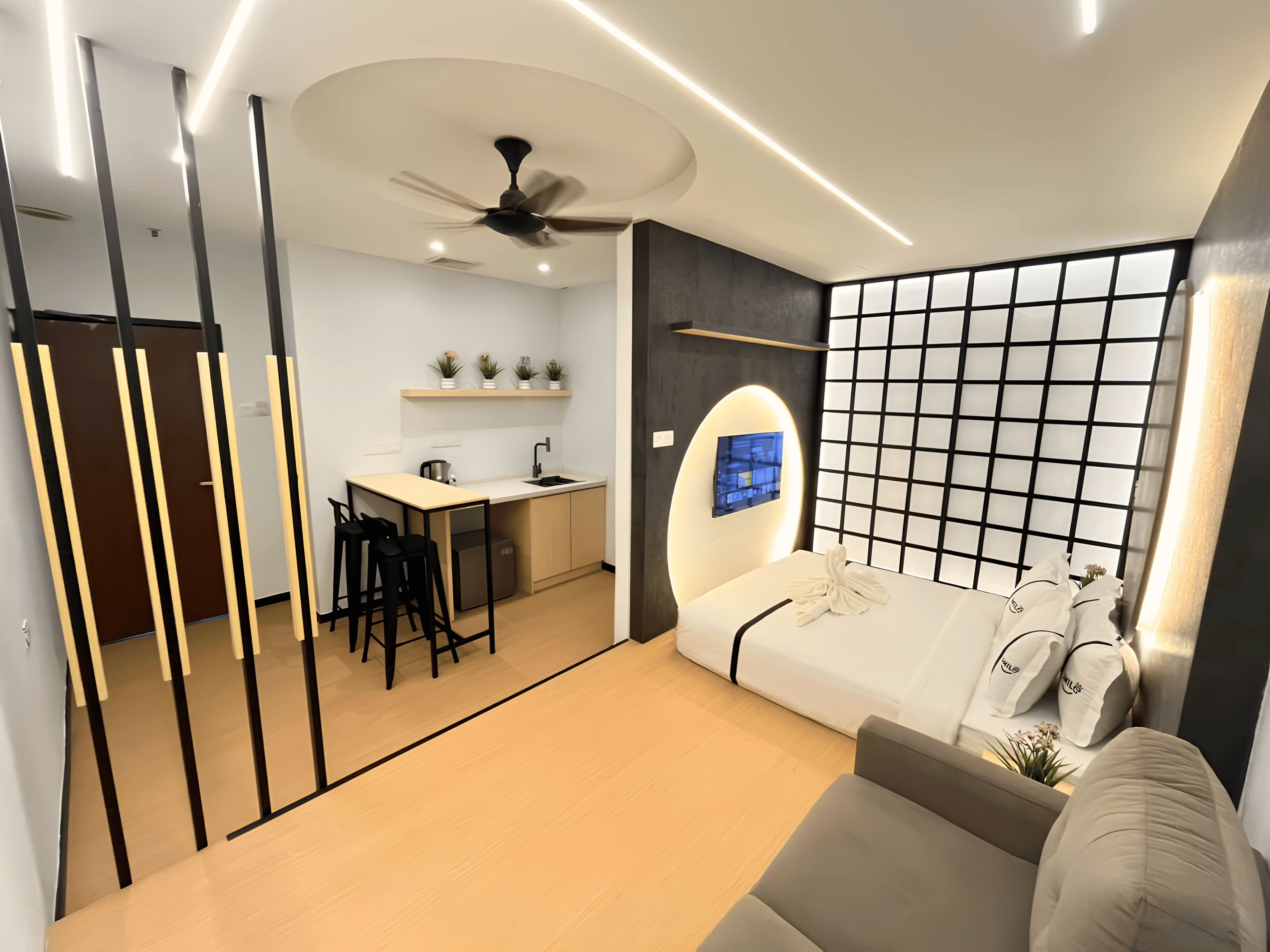 Ein kompakter Wohnbereich mit Küchenzeile, einem Bett mit Kissen, einem Deckenventilator und einem an der Wand befestigten Fernseher.