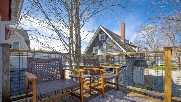 Une terrasse en bois avec deux fauteuils en osier, une clôture en métal et une vue sur une maison dotée d’une cheminée.