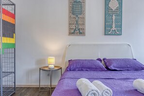 A bedroom with a bed, pillows, a nightstand with a lamp, and a wall with decorative items.
